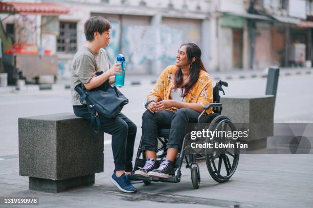 femme asiatique chinoise de mi-adulte parlant à son ami féminin indien avec le handicap sur le fauteuil roulant au trottoir de rue de ville - accessibilité aux personnes handicapées photos et images de collection