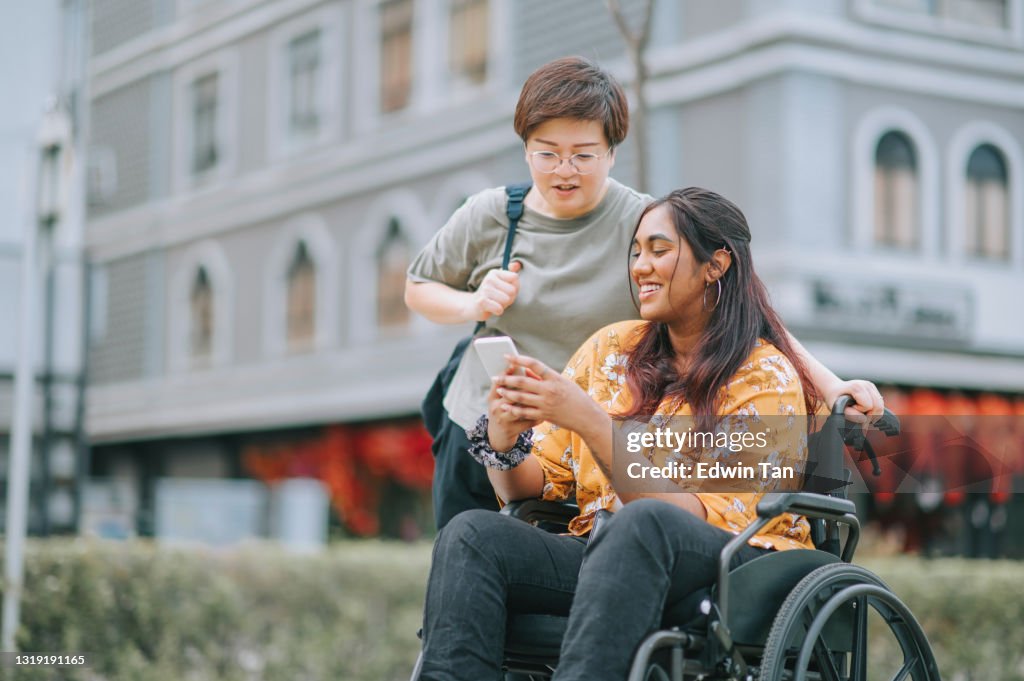 Tourist asian indian woman with wheelchair talking to her female chinese friend at sidewalk of the city