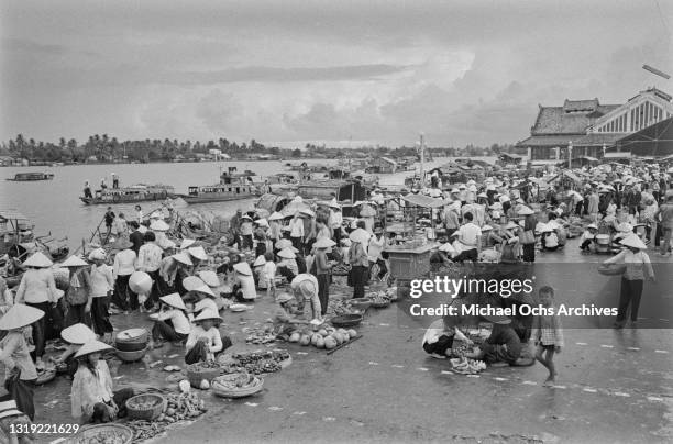 Vietnamese women, wearing traditional non la hats, selling fresh produce on an embankment of the Saigon River as it flows through Saigon, Vietnam,...