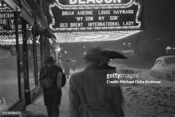 Pedestrians some carrying umbrellas as they walk along the snow-covered sidewalk outside the Beacon Theatre on Broadway in New York City, New York,...