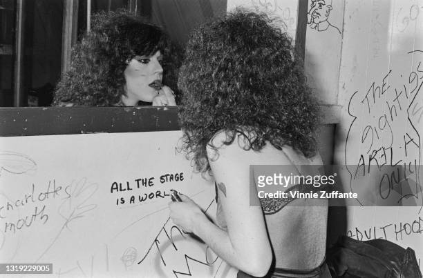 Woman with a loose perm applying lipstick while looking in a mirror which is fixed to a graffiti-strewn wall, location unspecified, March 1980. Among...