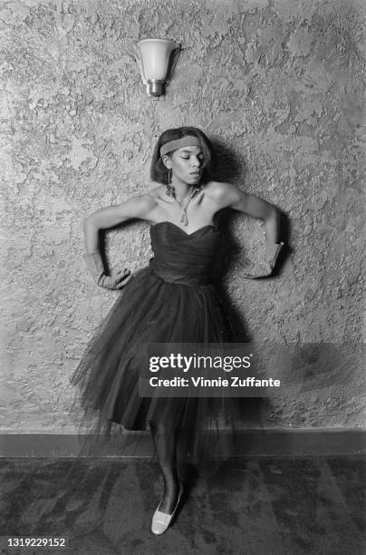 Young person wearing a dress with a tulle skirt and a sweetheart neckline, with a headband and evening gloves, poses with both arms against a...