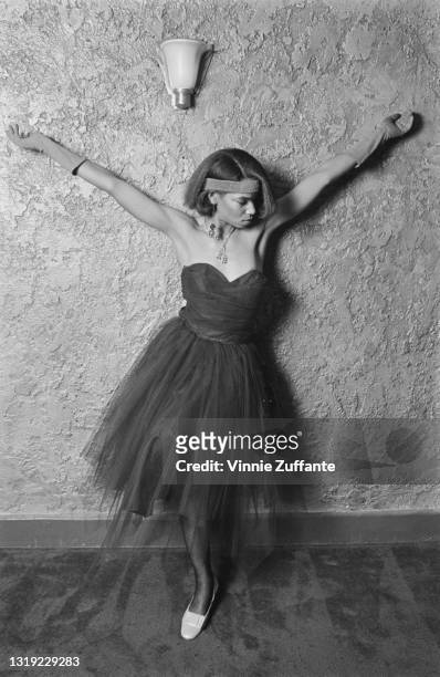 Young person wearing a dress with a tulle skirt and a sweetheart neckline, with a headband and evening gloves, poses with both arms raised against a...