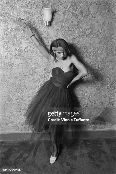 Young person wearing a dress with a tulle skirt and a sweetheart neckline, with a headband and evening gloves, poses with one arm raised, the other...