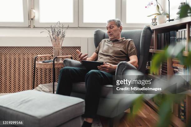 ein älterer mann sitzt in einem sessel und hat einen videoanruf mit einem arzt - dreiviertelansicht stock-fotos und bilder