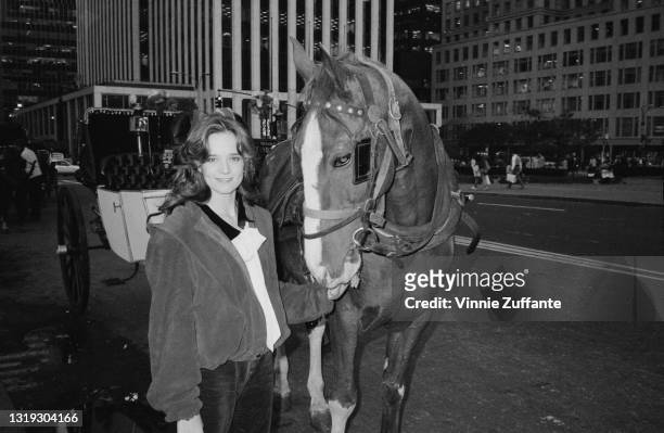 American actress Lea Thompson, wearing a blouson-syle jacket, poses with a horse and carriage with the facades of buildings and pedestrians in the...