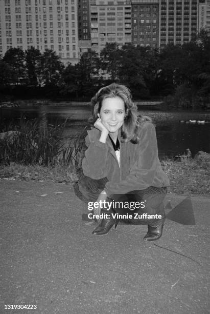 American actress Lea Thompson, wearing a blouson-syle jacket, crouches down with her chin resting in her hand, in the background is a lake and the...