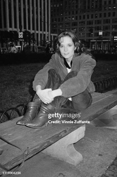 American actress Lea Thompson, wearing a blouson-syle jacket as she sits on a park bench, her arms around her knees, location unspecified, circa 1985.