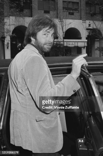 American actor and comedian John Ritter , unshaven and wearing a sports jacket over a sweater, stands before a car, location unspecified, circa 1985.