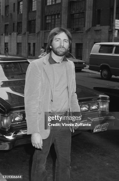 American actor and comedian John Ritter , unshaven and wearing a sports jacket over a sweater, stands before a car, location unspecified, circa 1985.