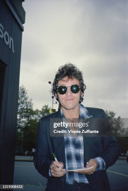American singer-songwriter and musician Bruce Springsteen, wearing sunglasses, a dark blue jacket over a blue checked shirt, holding a pad and pen,...