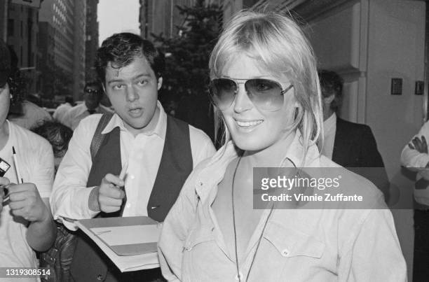 American actress Bo Derek, wearing gradually tinted sunglasses and a blouse with the collar open, smiling alongside what appears to be a journalist...