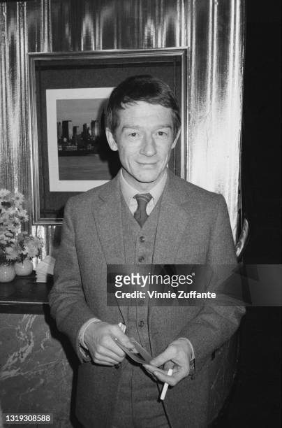 British-born American actor Roddy McDowall , wearing a three-piece suit, with a cigarette in his hand and a framed picture on the wall behind him,...