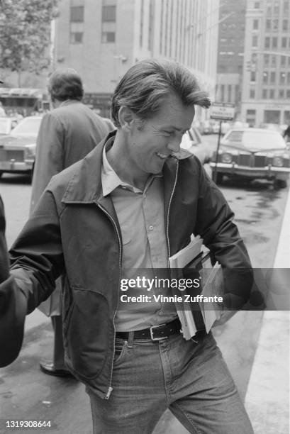 American actor Martin Sheen, wearing a light jacket over a shirt with the collar open and jeans, smiling as carries books and documents under his...