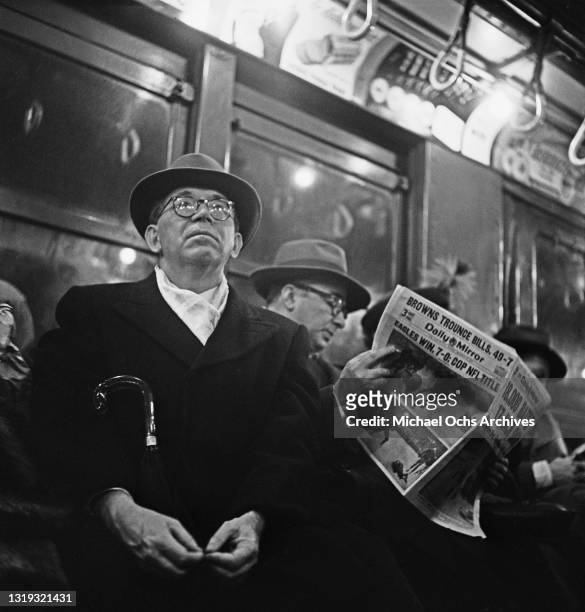 Man in an overcoat and hat, his umbrella under his arm sits alongside a man reading a newspaper with the headline on the sports pages reading 'Eagles...