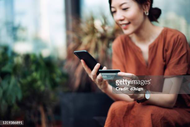 beautiful smiling young asian woman relaxing on deck chair in the backyard, surrounded by beautiful houseplants. shopping online on smartphone and making mobile payment with credit card. technology makes life so much easier. lifestyle and technology - credit card stock-fotos und bilder