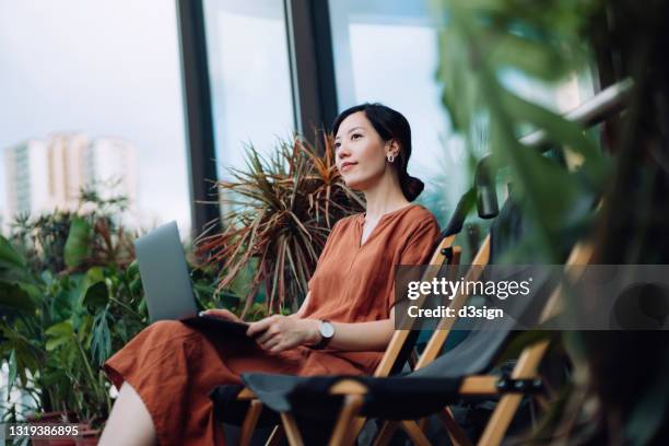 contemplated young asian woman looking away in thought while relaxing on deck chair using laptop in the backyard, surrounded by beautiful houseplants. lifestyle and technology - einfaches leben stock-fotos und bilder