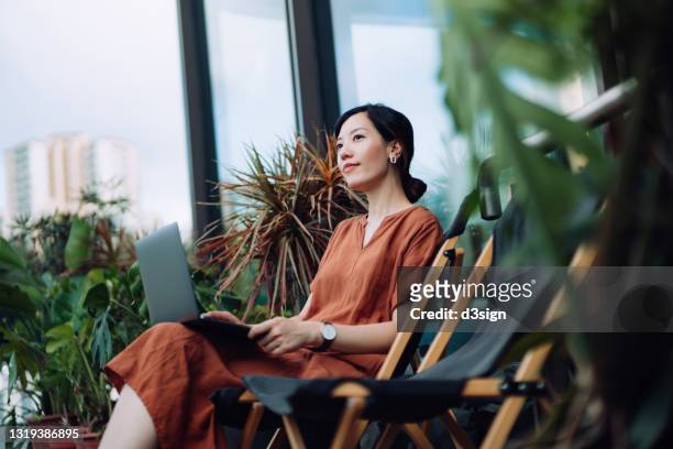 contemplated young asian woman looking away in thought while relaxing on deck chair using laptop in the backyard, surrounded by beautiful houseplants. lifestyle and technology - real estate sign fotografías e imágenes de stock