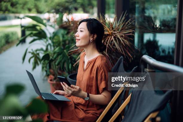 contemplated young asian woman looking away in thought while using smartphone and working on laptop. relaxing on deck chair in the backyard, surrounded by beautiful houseplants. lifestyle and technology - real estate sign fotografías e imágenes de stock
