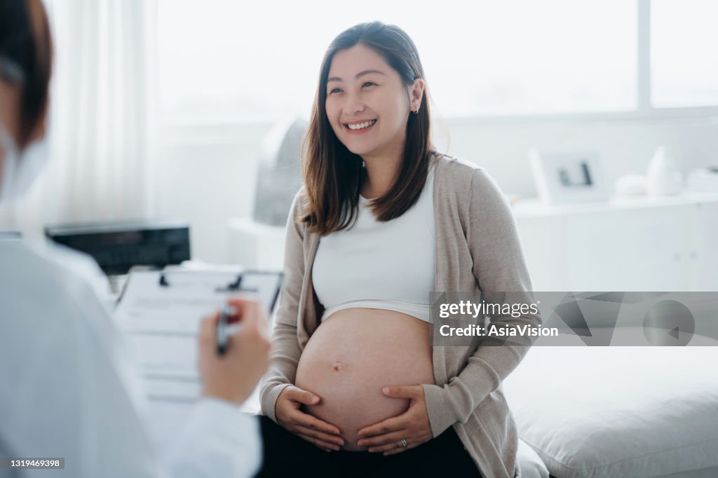 Leende ung asiatisk gravid kvinna som har en konsultation med en kvinnlig läkare under hennes rutinkontroll på en klinik. Kontroll, tester och skanningar för att säkerställa en hälsosam graviditet för både mamma och det ofödda barnet. Hälsosam gr