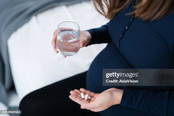 cierre la sección media de la mujer embarazada asiática sentada en el lado de la cama en casa, tomando medicamentos en la mano con un vaso de agua. las vitaminas y suplementos esenciales son vitales para el crecimiento y desarrollo saludable del feto - calcita fotografías e imágenes de stock