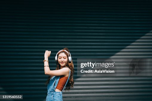 色の壁や日光に対して屋外のヘッドフォンで音楽を聴きながら、目を閉じて踊る気楽な笑顔の若いアジア人女性。音楽とライフスタイル 色の壁や日光に対して屋外のヘッドフォンで音楽を聴きながら、目を閉じて踊る気楽な笑顔の若いアジア人女性。音楽とライフスタイル