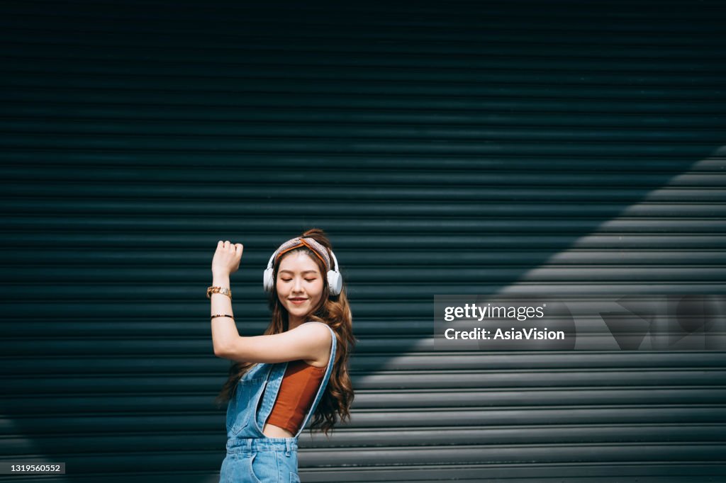 無憂無慮的微笑的亞洲年輕女子閉著眼睛跳舞,同時在戶外的耳機上聽音樂,面對著彩色的牆壁和陽光。音樂和生活方式