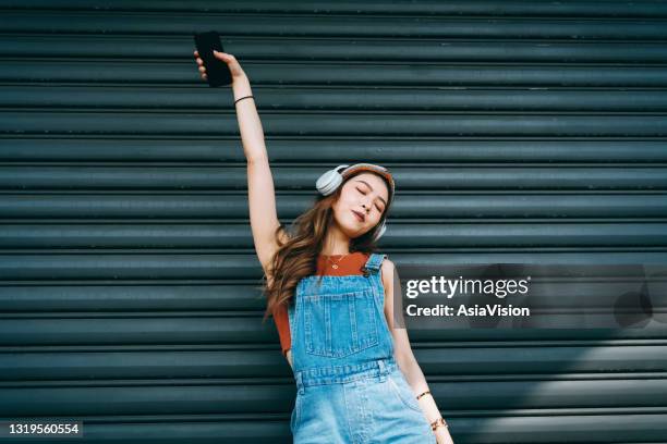 carefree smiling young asian woman holding smartphone, dancing with her eyes closed while listening to music on headphones outdoors against coloured wall and sunlight. music and lifestyle - call to arms stock pictures, royalty-free photos & images