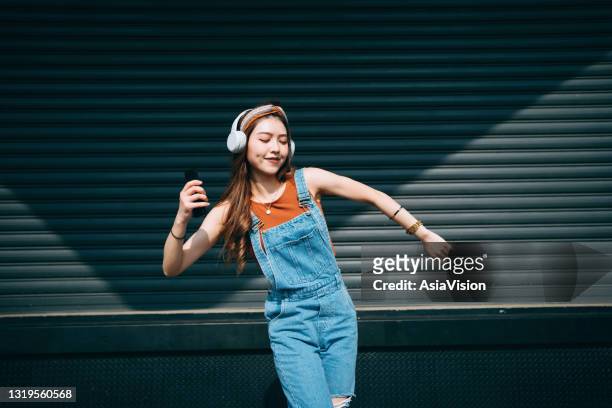 スマートフォンを持つ気楽な笑顔の若いアジア人女性は、色の壁や日光に対して屋外のヘッドフォンで音楽を聴きながら目を閉じて踊ります。音楽とライフスタイル - very young asian girls ストックフォトと画像