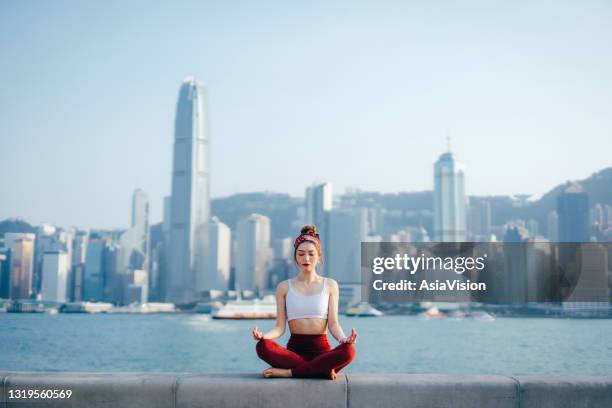 junge asiatische sportlerin meditiert im freien, übt achtsamkeit yoga am morgen, gegen spektakuläre hong kong stadt skyline an der promenade von victoria hafen. girl power, wohlbefinden, fitness und gesundes lifestyle-konzept - holy city park stock-fotos und bilder