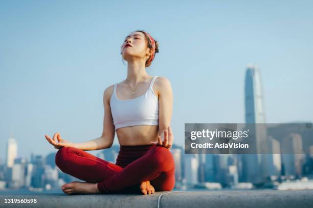 young asian sports woman meditating outdoors, practicing mindfulness yoga in the morning, against spectacular hong kong city skyline by the promenade of victoria harbour. girl power, wellbeing, fitness and healthy lifestyle concept - commercial buildings hong kong morning stock pictures, royalty-free photos & images