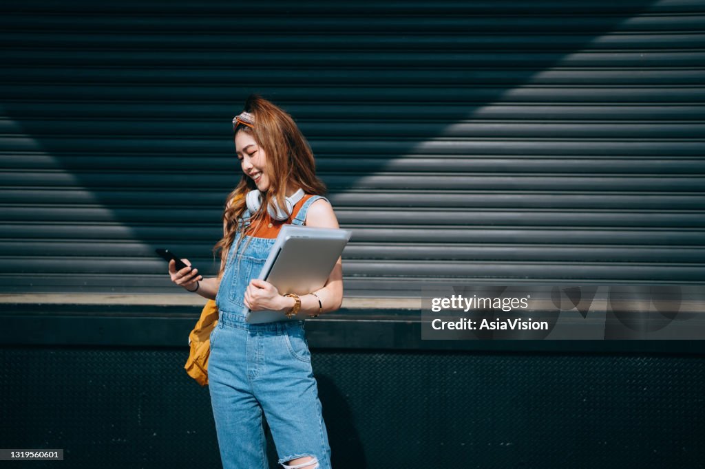 Smiling female university student carrying backpack and wearing headphones around her neck. She is text messaging on smartphone and holding laptop while walking in school campus against sunlight. Teenage lifestyle and technology
