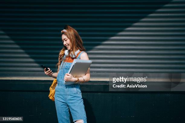 smiling female university student carrying backpack and wearing headphones around her neck. she is text messaging on smartphone and holding laptop while walking in school campus against sunlight. teenage lifestyle and technology - beautiful college girls stock pictures, royalty-free photos & images