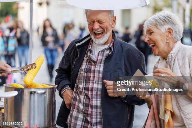 una coppia più anziana sta mangiando una pannocchia di mais bollita e camminando in città. - bbq corn foto e immagini stock