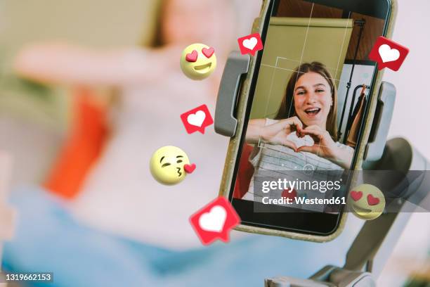teenage female influencer showing heart shape while filming through mobile phone with social media icons around at home - excitement emoji stock pictures, royalty-free photos & images
