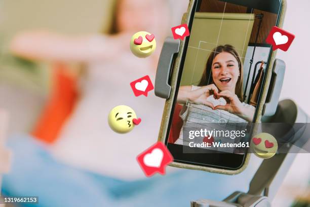 teenage female influencer showing heart shape while filming through mobile phone with social media icons around at home - teen doing filming imagens e fotografias de stock