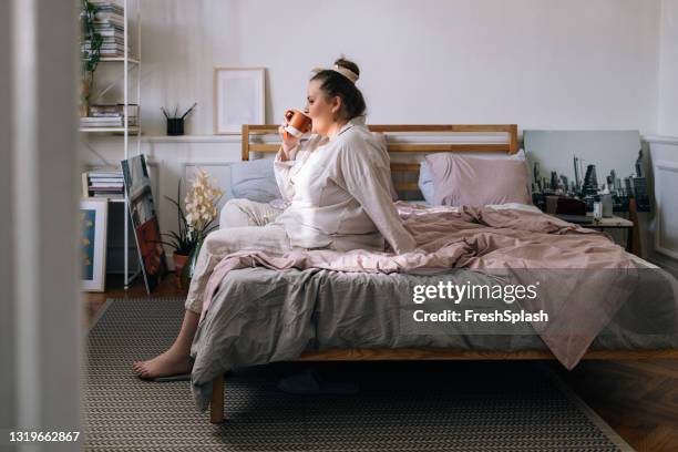 de zitting van de vrouw van het overgewicht op haar bed en het drinken van thee - slaapkamer zijaanzicht stockfoto's en -beelden