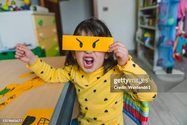 girl making angry face while holding emoticon at home - aggressive stock-fotos und bilder