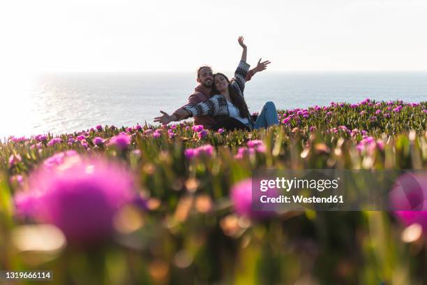 young couple with arms outstretched sitting at flower filed by sea - barrilha imagens e fotografias de stock