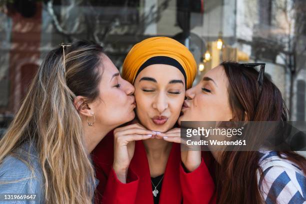 female friends kissing young woman wearing headscarf outdoors - funny face stock pictures, royalty-free photos & images