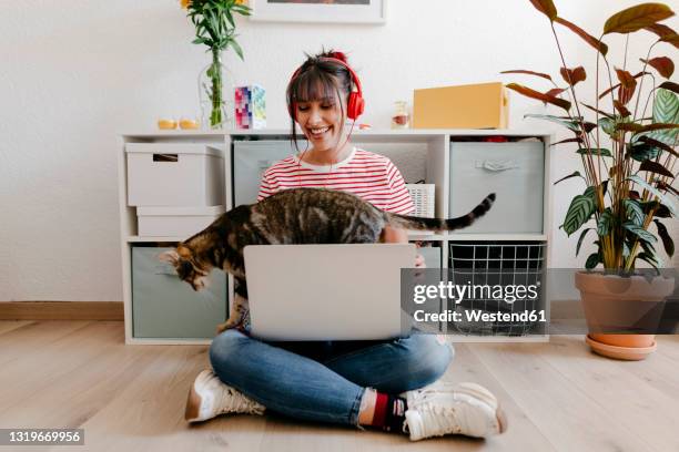 young woman wearing headphones sitting with laptop and cat at home - happy millennial at home photos et images de collection