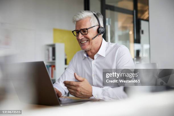 smiling businessman with headset gesturing while talking to video call on laptop in office - man on video call stock pictures, royalty-free photos & images