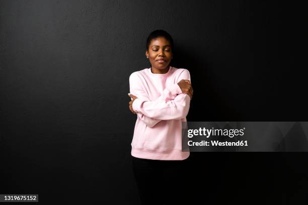 smiling woman wearing sweatshirt against black background - hugging self stock-fotos und bilder