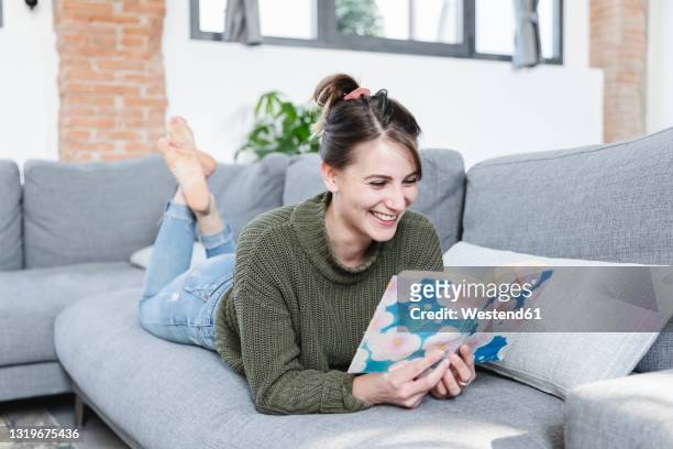 smiling woman reading book while lying on sofa at home - china foto e immagini stock