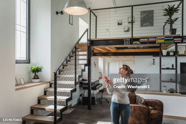young woman controlling smart home functions through mobile phone at modern home - connected home fotografías e imágenes de stock