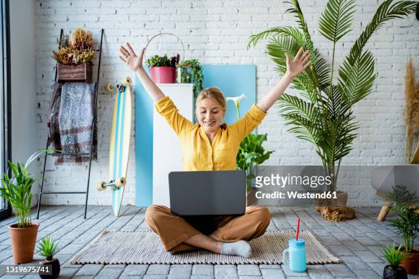 businesswoman with arms raised sitting with laptop at home - 胡坐　横 ストックフォトと画像