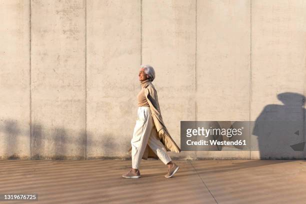 mature woman in trench coat walking on footpath - fashion stock-fotos und bilder