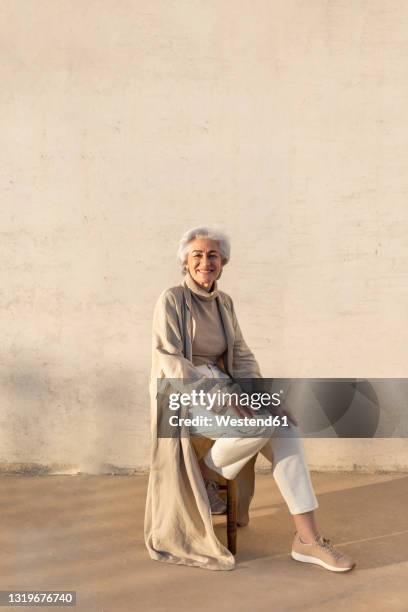 smiling woman sitting on stool by wall - old woman sitting stock-fotos und bilder