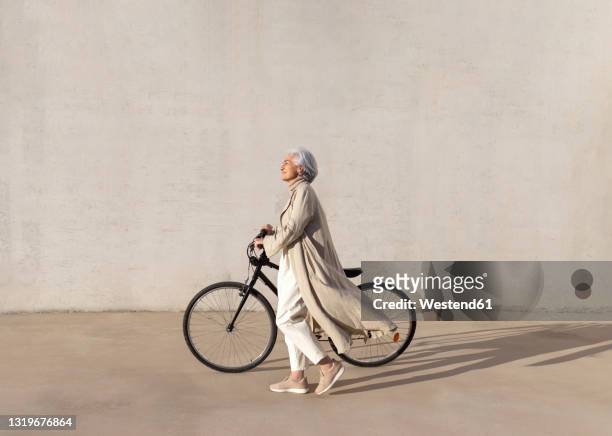 mature woman walking with bicycle on footpath during sunny day - cabello gris fotografías e imágenes de stock