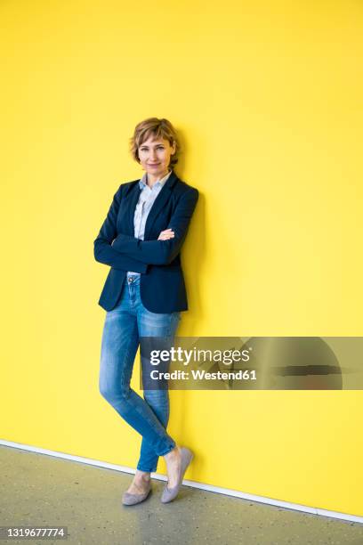 confident businesswoman with arms crossed leaning on yellow wall - veste jaune photos et images de collection